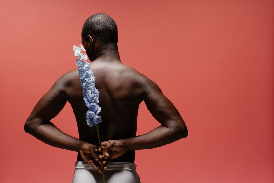 A shirtless man holding a blue flower against a pink background.
