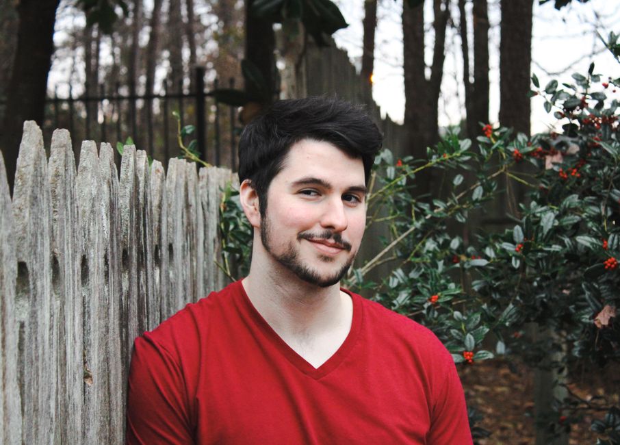 Portrait of a smiling man in a red shirt leaning against a wooden fence outdoors.